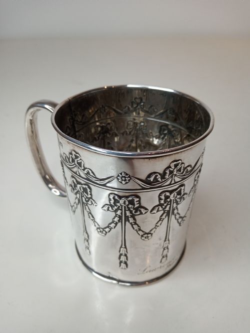 fig 1. An Edwardian sterling silver christening mug featuring chased floral swags, ribbon bow motifs, and a smooth loop handle.