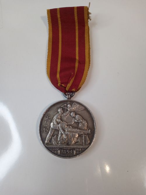fig 1. An antique silver circular service medal dated 1894 featuring relief figures and Chinese characters, suspended from a red and yellow striped silk ribbon.