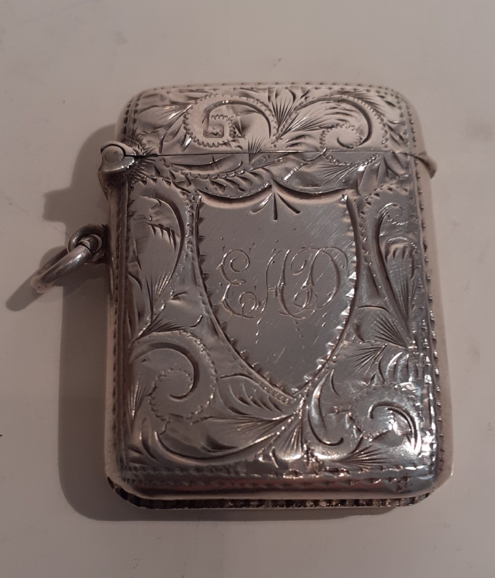 An Edwardian sterling silver vesta case featuring hand-engraved foliate scrollwork, a central monogrammed shield cartouche, a side suspension ring, and a hinged lid.