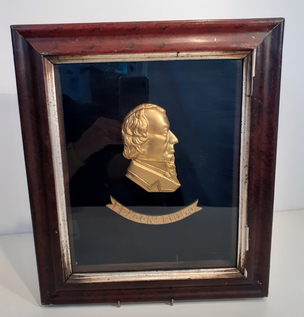 A framed Victorian profile relief portrait of Benjamin Disraeli in gold-coloured brass, inscribed with Beaconsfield, set against a black background inside a mahogany-stained wood frame.