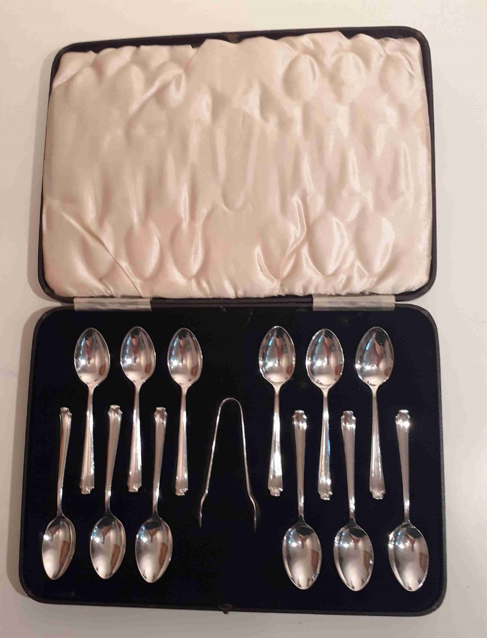 A cased set of twelve silver teaspoons and matching sugar tongs arranged in a dark velvet-lined presentation box.