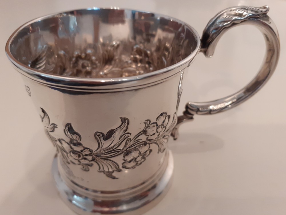 A Victorian silver christening mug featuring floral embossed scrollwork, a flared rim, and a curved leaf-capped handle on a circular pedestal base.