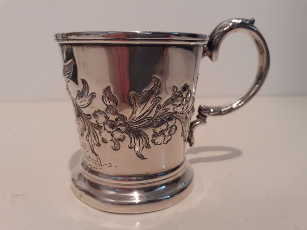 A Victorian silver christening mug featuring floral embossed scrollwork, a flared rim, and a curved leaf-capped handle on a circular pedestal base. View II