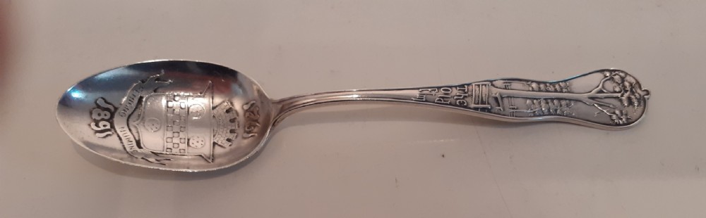 An antique silver souvenir spoon featuring an embossed heraldic crest in the bowl dated 1891 and a decorative handle depicting an elm tree.