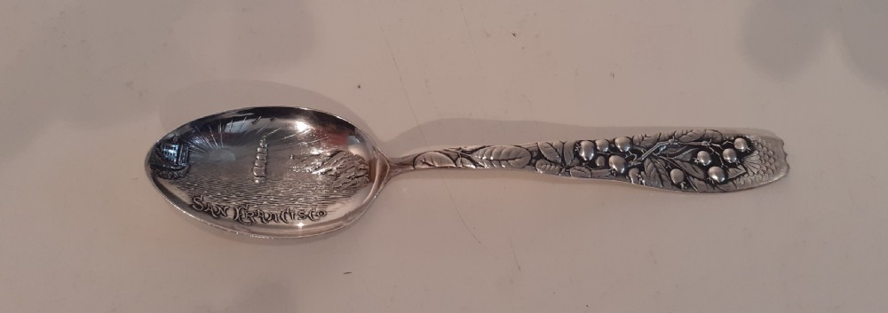 An antique silver souvenir spoon featuring an embossed depiction of San Francisco in the bowl and detailed botanical relief motifs on the handle.