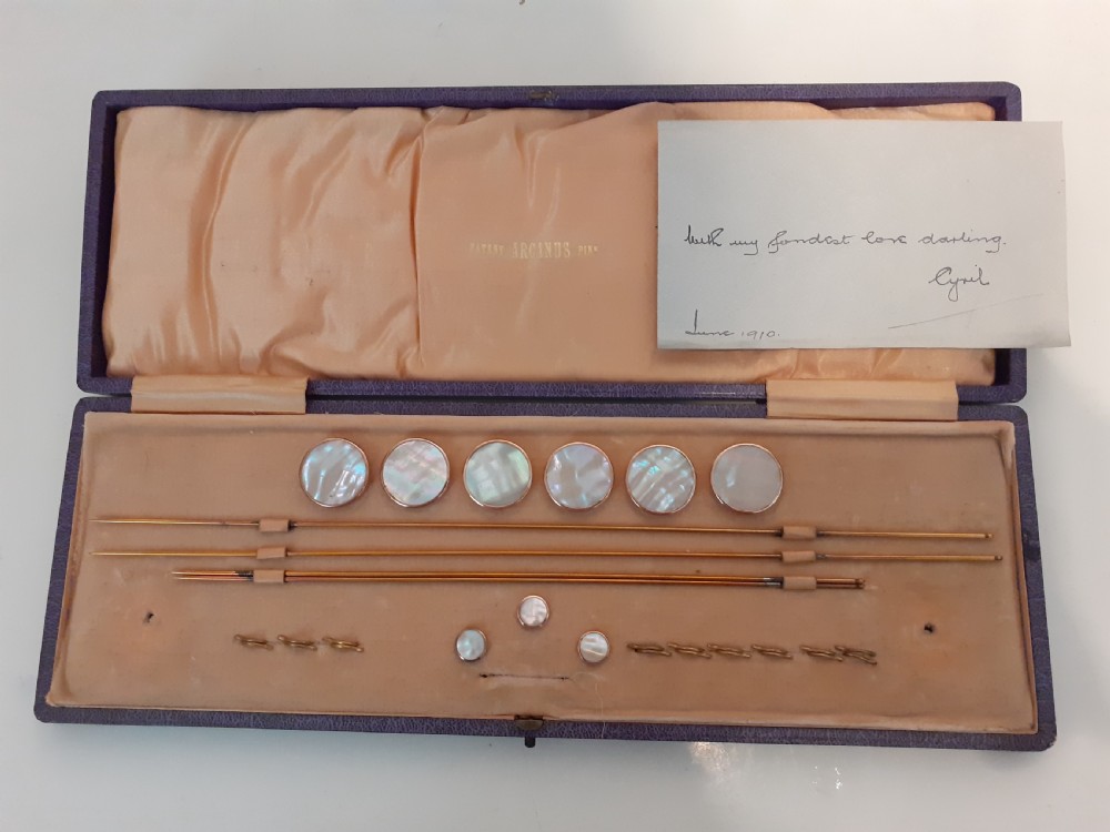 A boxed set of circular mother of pearl dress studs, pins, and metal fittings presented inside a purple satin-lined case.