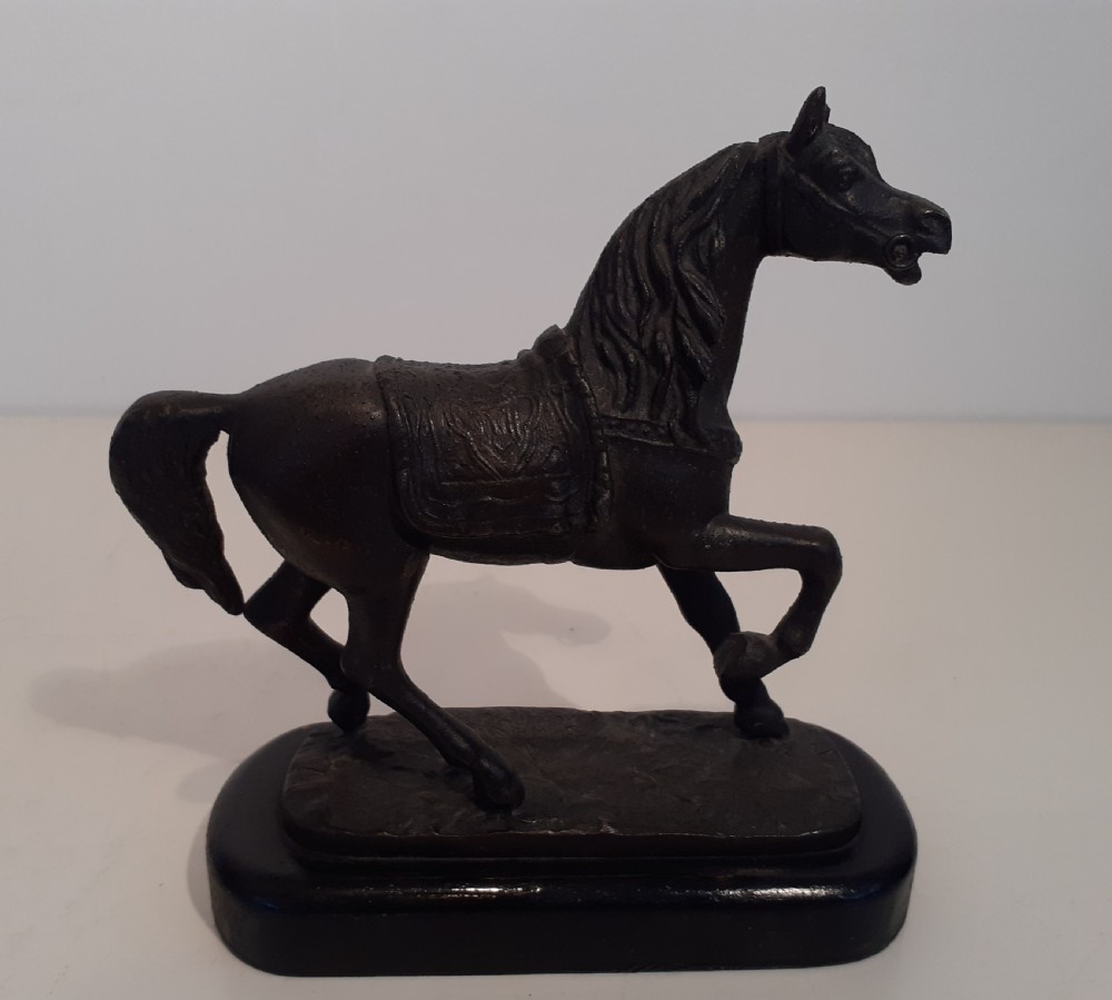 A dark cast metal model of a horse in a trotting pose with saddle and bridle, mounted on an oval dark wood base.