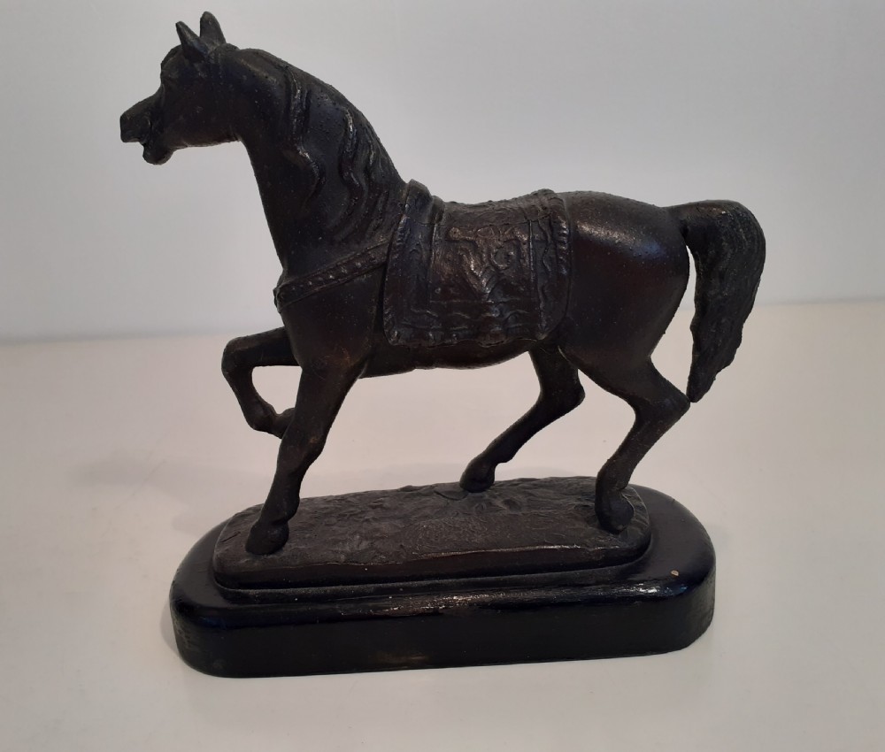 A dark cast metal model of a horse in a trotting pose with saddle and bridle, mounted on an oval dark wood base. View II