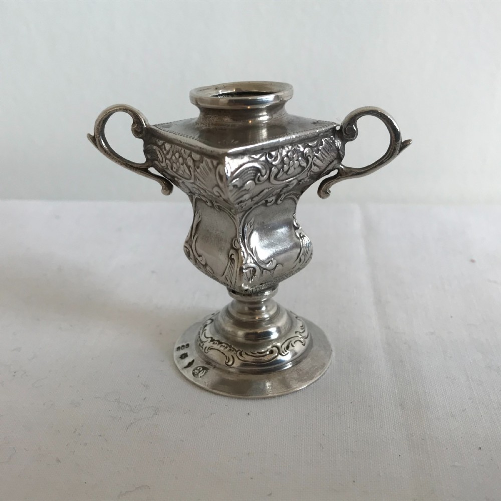 An ornate German silver miniature urn vase featuring scrolled handles, floral relief chasing, and a circular pedestal base.