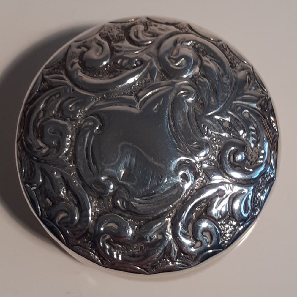 A circular antique sterling silver pill box featuring raised scrollwork foliage and an unengraved central cartouche.