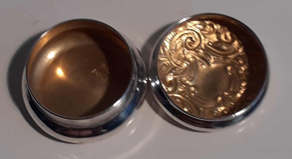 A circular antique sterling silver pill box featuring raised scrollwork foliage and an unengraved central cartouche. View II