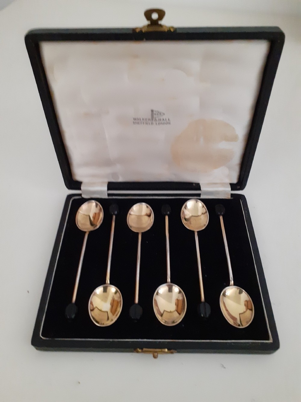 A set of six silver gilt coffee spoons with dark bean finials resting inside an open fitted presentation case lined with cream satin and dark velvet.