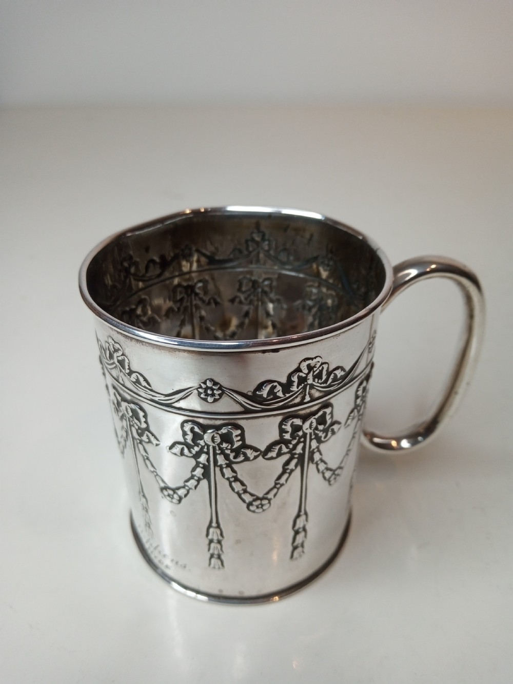 An Edwardian sterling silver christening mug featuring chased floral swags, ribbon bow motifs, and a smooth loop handle.