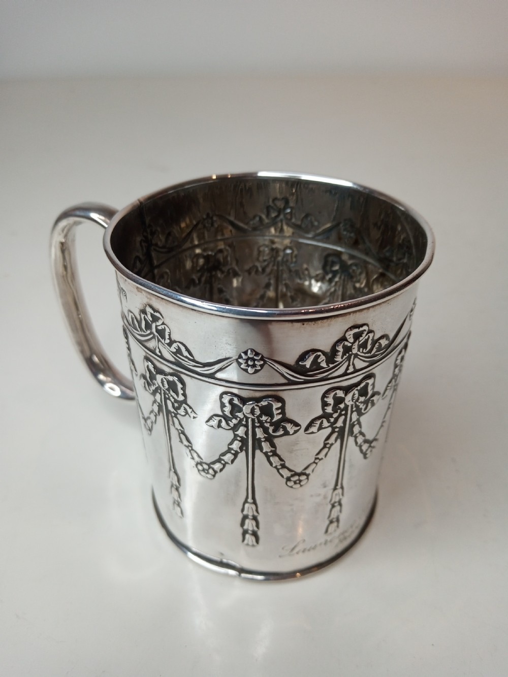 An Edwardian sterling silver christening mug featuring chased floral swags, ribbon bow motifs, and a smooth loop handle. View II