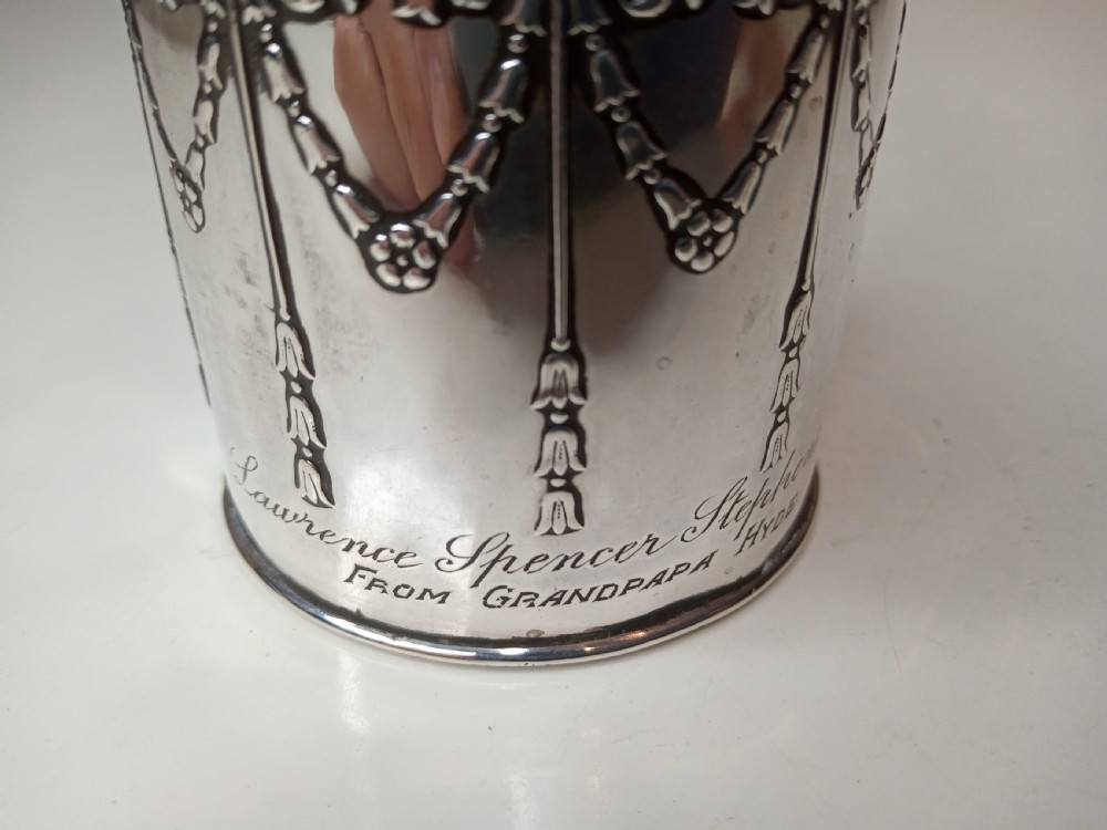 An Edwardian sterling silver christening mug featuring chased floral swags, ribbon bow motifs, and a smooth loop handle. View III