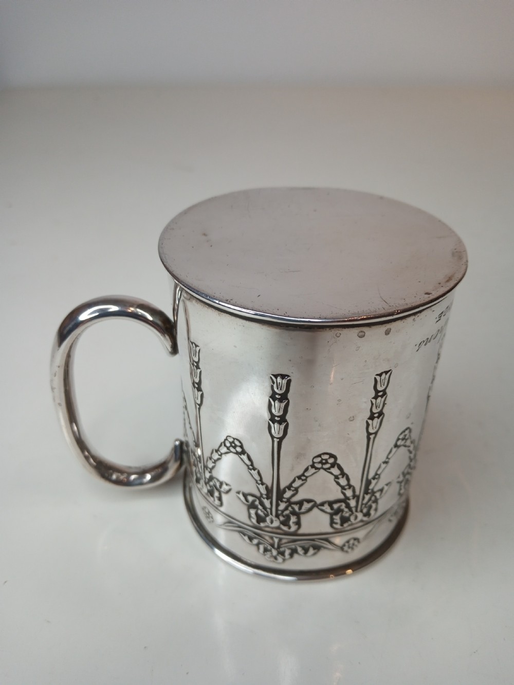 An Edwardian sterling silver christening mug featuring chased floral swags, ribbon bow motifs, and a smooth loop handle. View IV