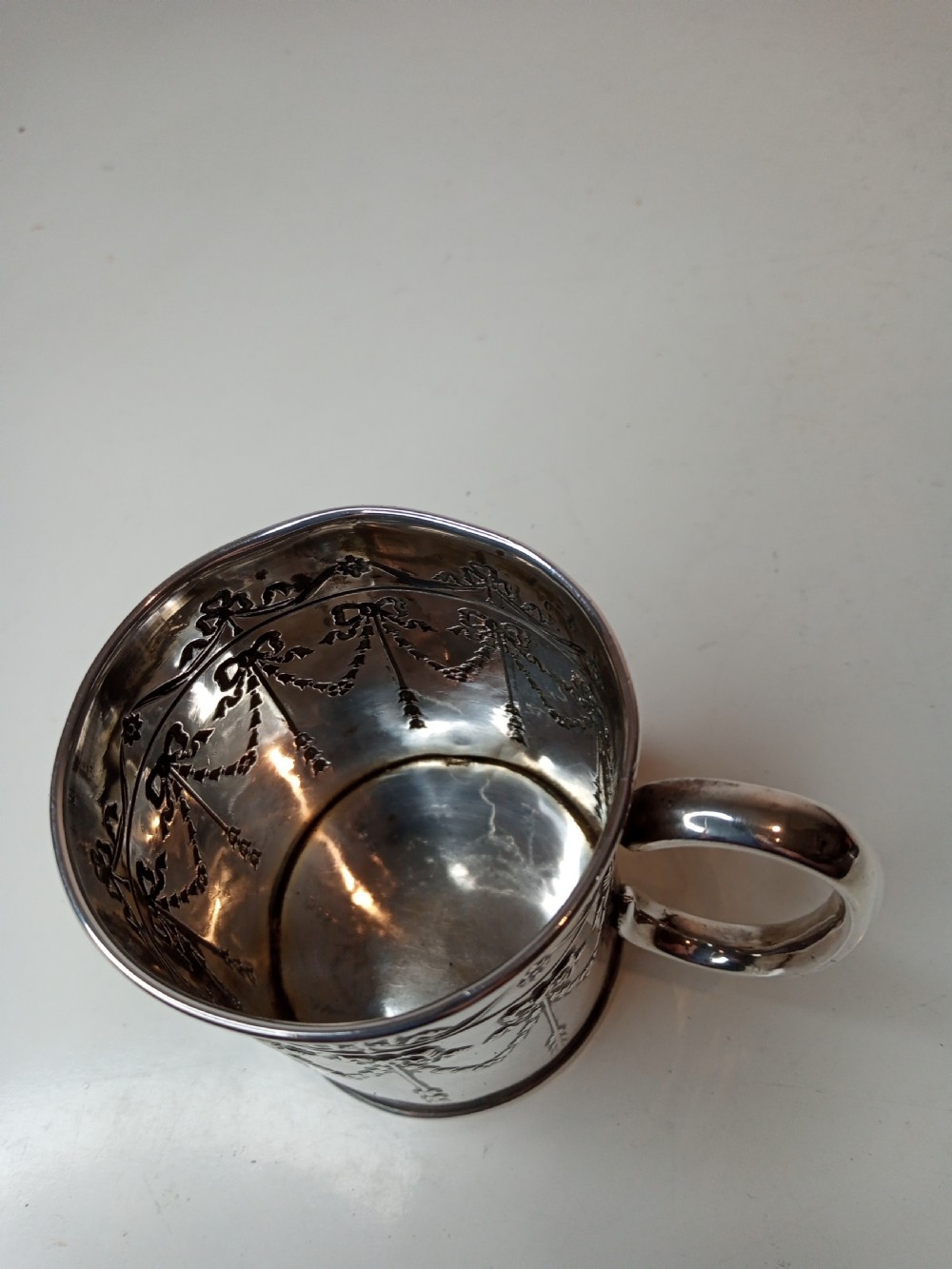An Edwardian sterling silver christening mug featuring chased floral swags, ribbon bow motifs, and a smooth loop handle. View V