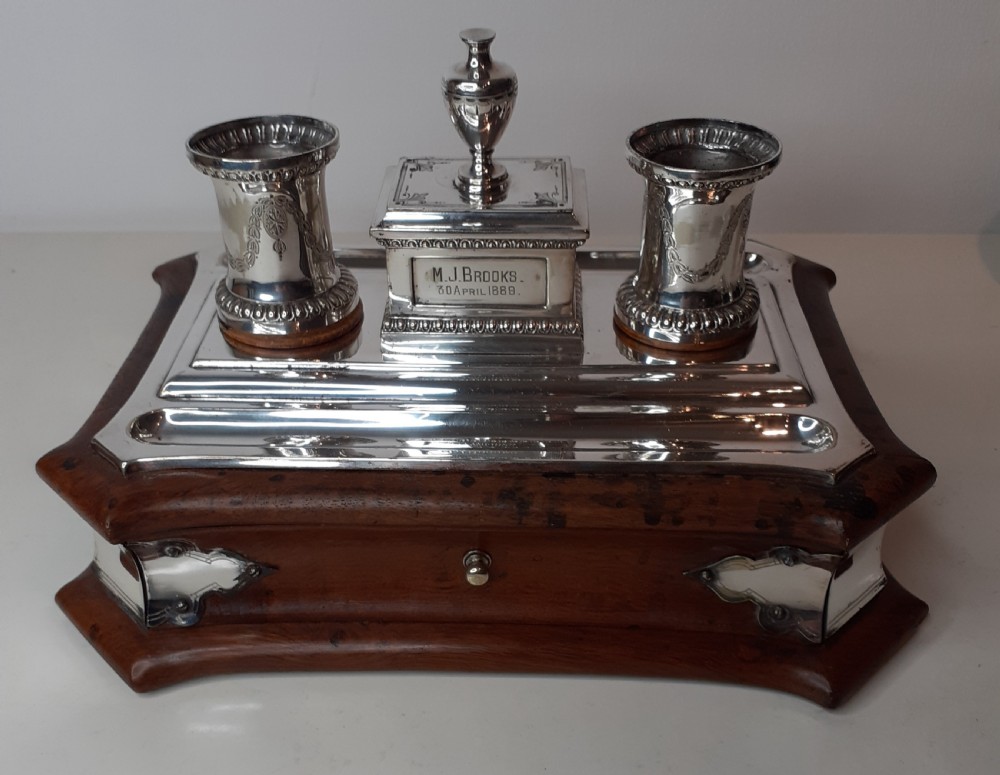 A Victorian silverplated standish featuring a shaped wooden base, two etched cylindrical inkwell holders, a central urn-topped box, and an integrated pen tray.