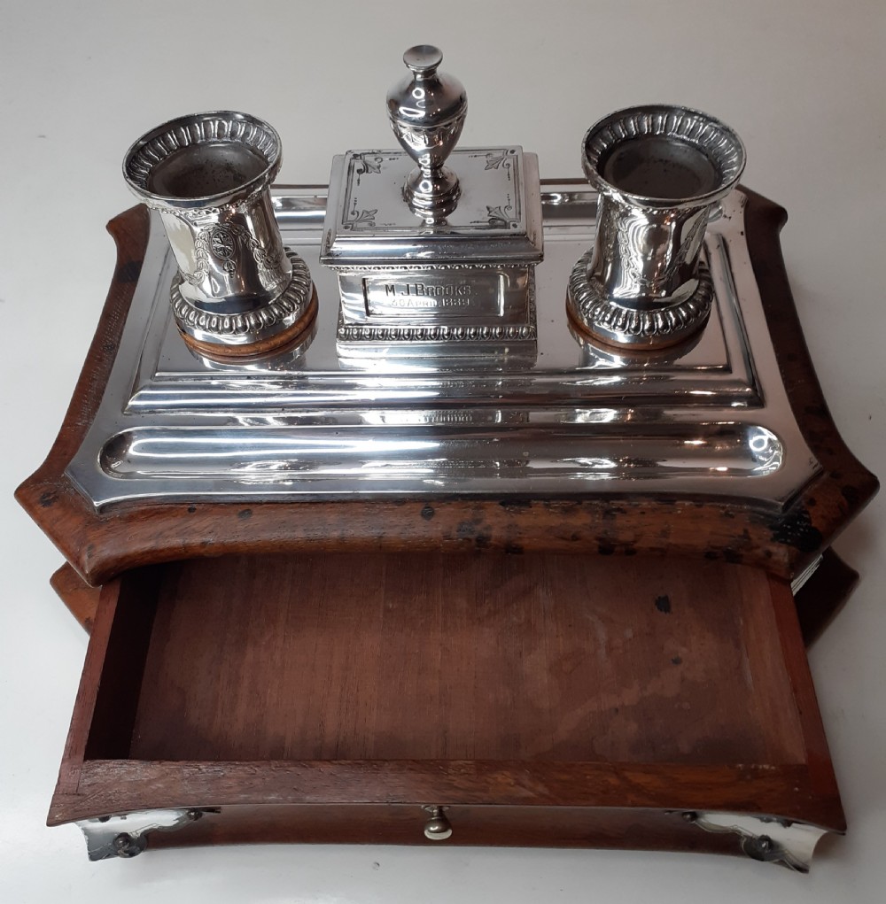 A Victorian silverplated standish featuring a shaped wooden base, two etched cylindrical inkwell holders, a central urn-topped box, and an integrated pen tray. View III
