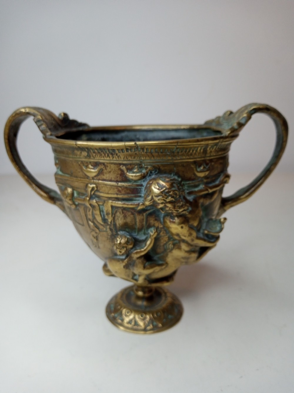 An antique brass kantharos cup featuring raised mythological relief figures, twin high-arched handles, and a circular pedestal base with green patinated oxidisation.