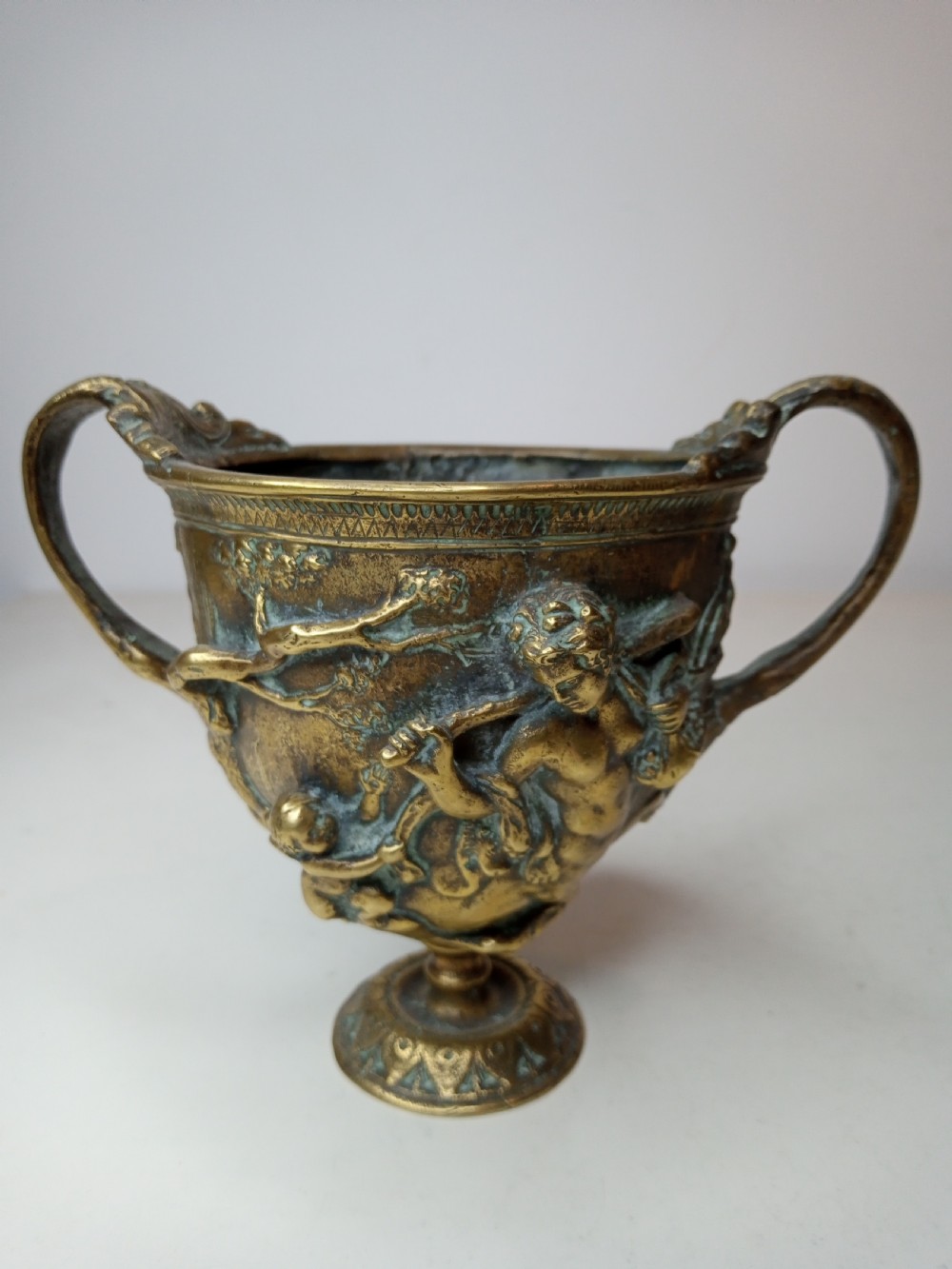 An antique brass kantharos cup featuring raised mythological relief figures, twin high-arched handles, and a circular pedestal base with green patinated oxidisation. View II