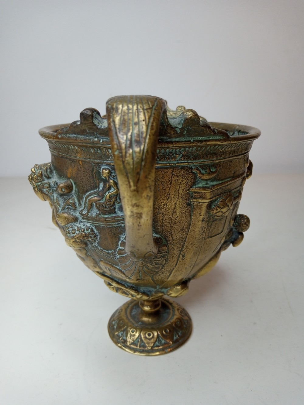 An antique brass kantharos cup featuring raised mythological relief figures, twin high-arched handles, and a circular pedestal base with green patinated oxidisation. View III