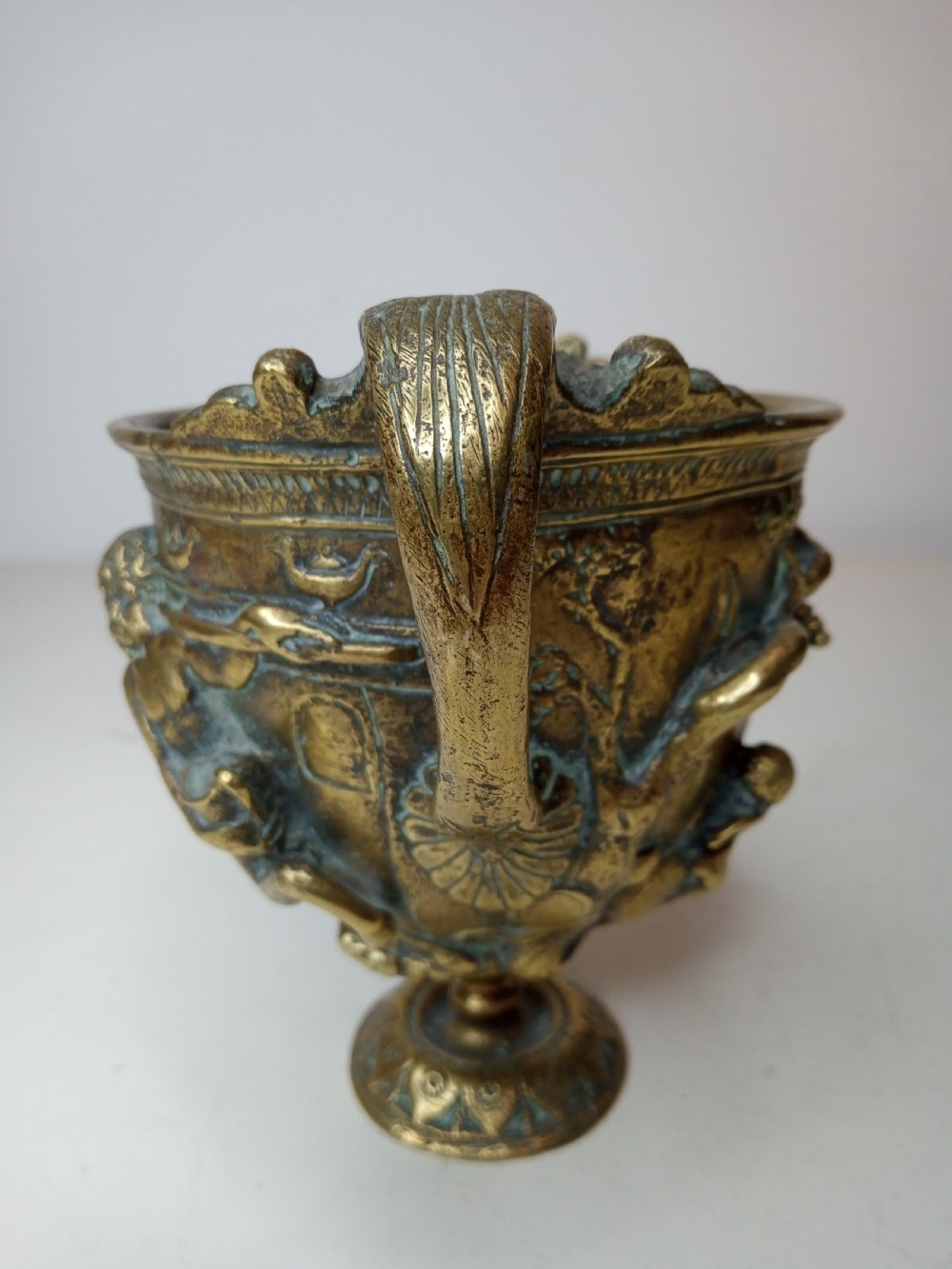 An antique brass kantharos cup featuring raised mythological relief figures, twin high-arched handles, and a circular pedestal base with green patinated oxidisation. View IV