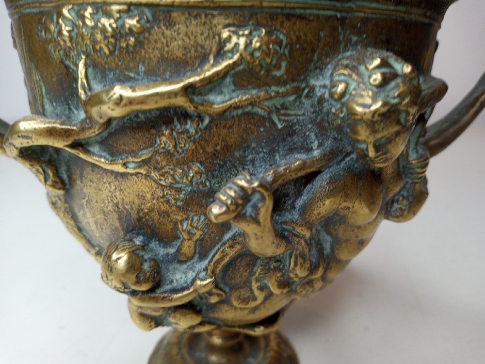 An antique brass kantharos cup featuring raised mythological relief figures, twin high-arched handles, and a circular pedestal base with green patinated oxidisation. View V