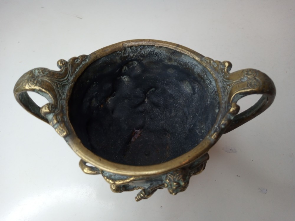 An antique brass kantharos cup featuring raised mythological relief figures, twin high-arched handles, and a circular pedestal base with green patinated oxidisation. View VI