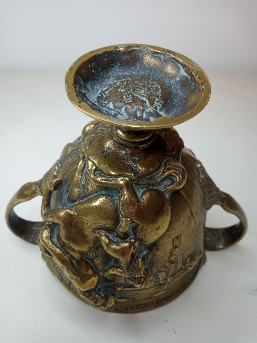 An antique brass kantharos cup featuring raised mythological relief figures, twin high-arched handles, and a circular pedestal base with green patinated oxidisation. View VII