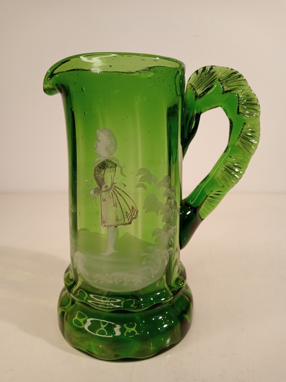 A Victorian emerald green glass pitcher featuring white enamel painting of a child figure among foliage, complete with a textured applied handle.