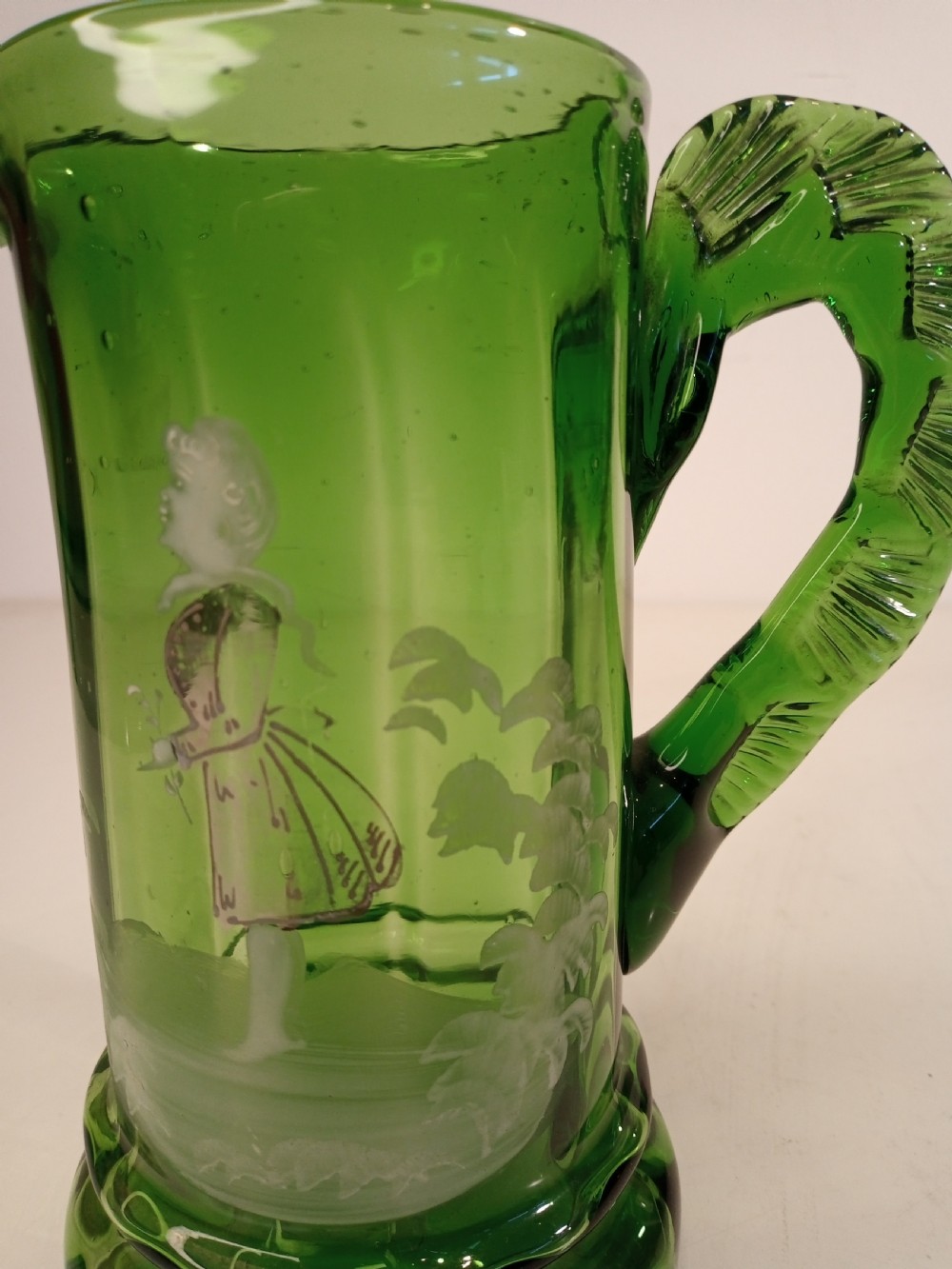A Victorian emerald green glass pitcher featuring white enamel painting of a child figure among foliage, complete with a textured applied handle. View V