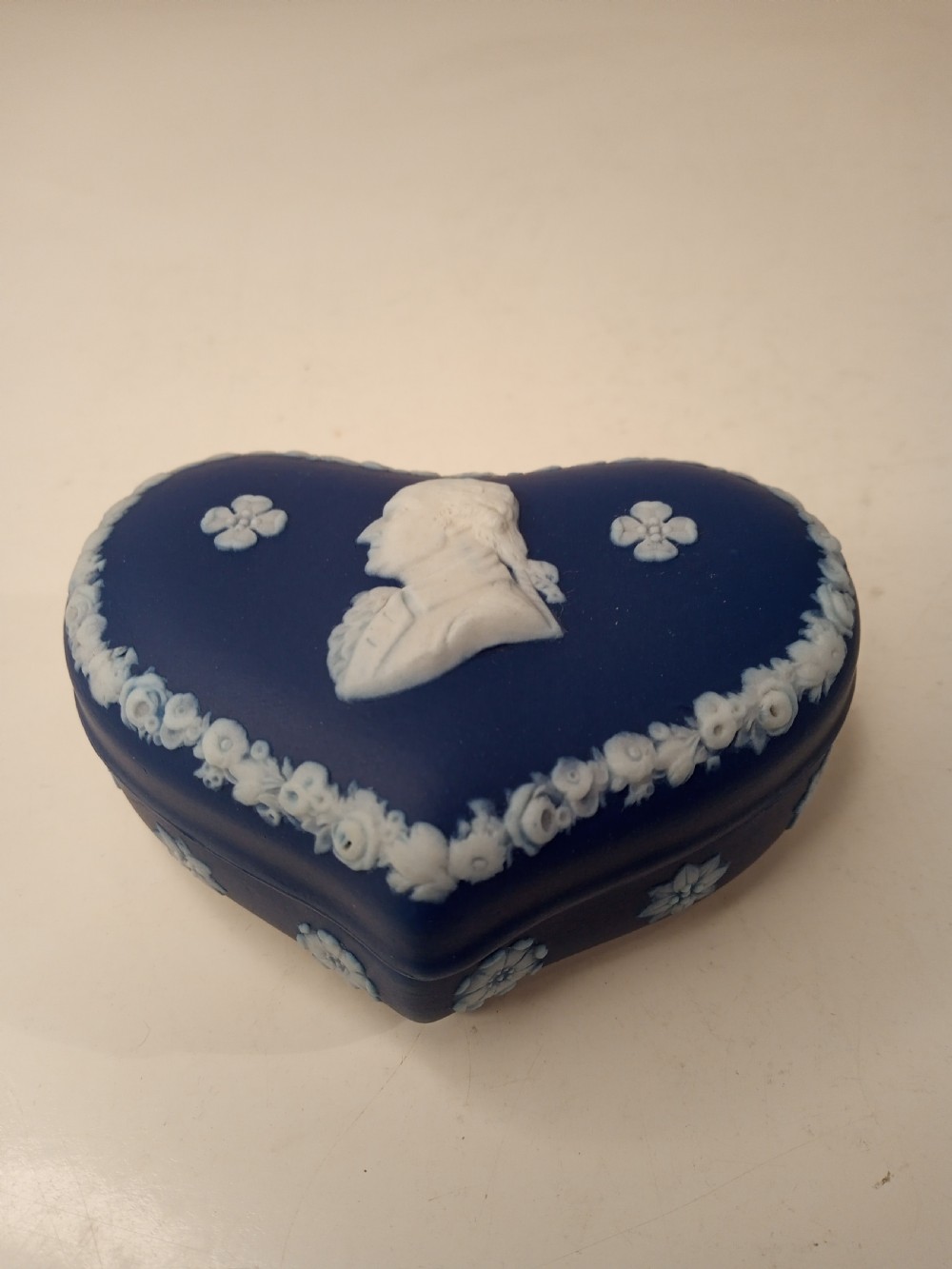 A heart-shaped dark blue Jasperware trinket box lid featuring a white bas-relief profile portrait bust surrounded by floral border accents. View II