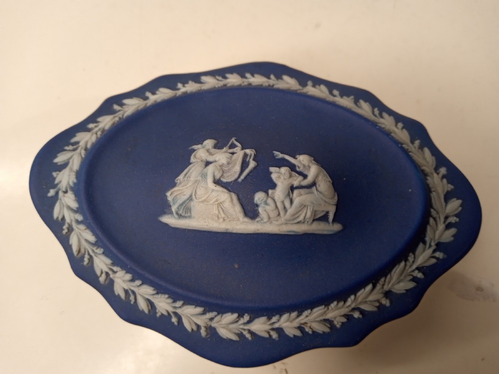 A dark blue oval jasperware lidded box featuring raised white classical bas-relief figures on the top surrounded by a white laurel leaf border.