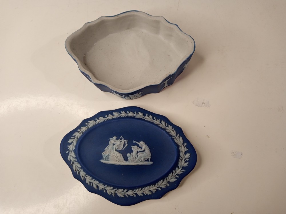 A dark blue oval jasperware lidded box featuring raised white classical bas-relief figures on the top surrounded by a white laurel leaf border. View II