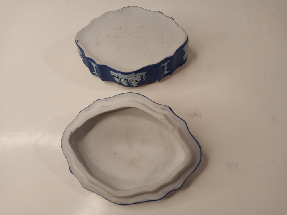 A dark blue oval jasperware lidded box featuring raised white classical bas-relief figures on the top surrounded by a white laurel leaf border. View III