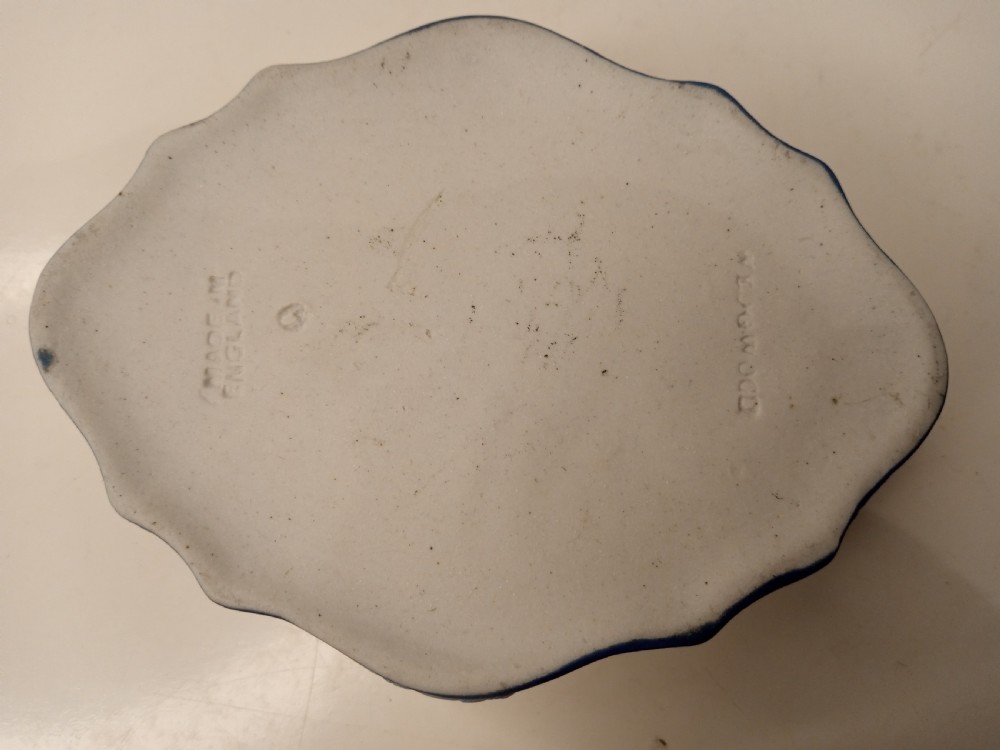 A dark blue oval jasperware lidded box featuring raised white classical bas-relief figures on the top surrounded by a white laurel leaf border. View IV