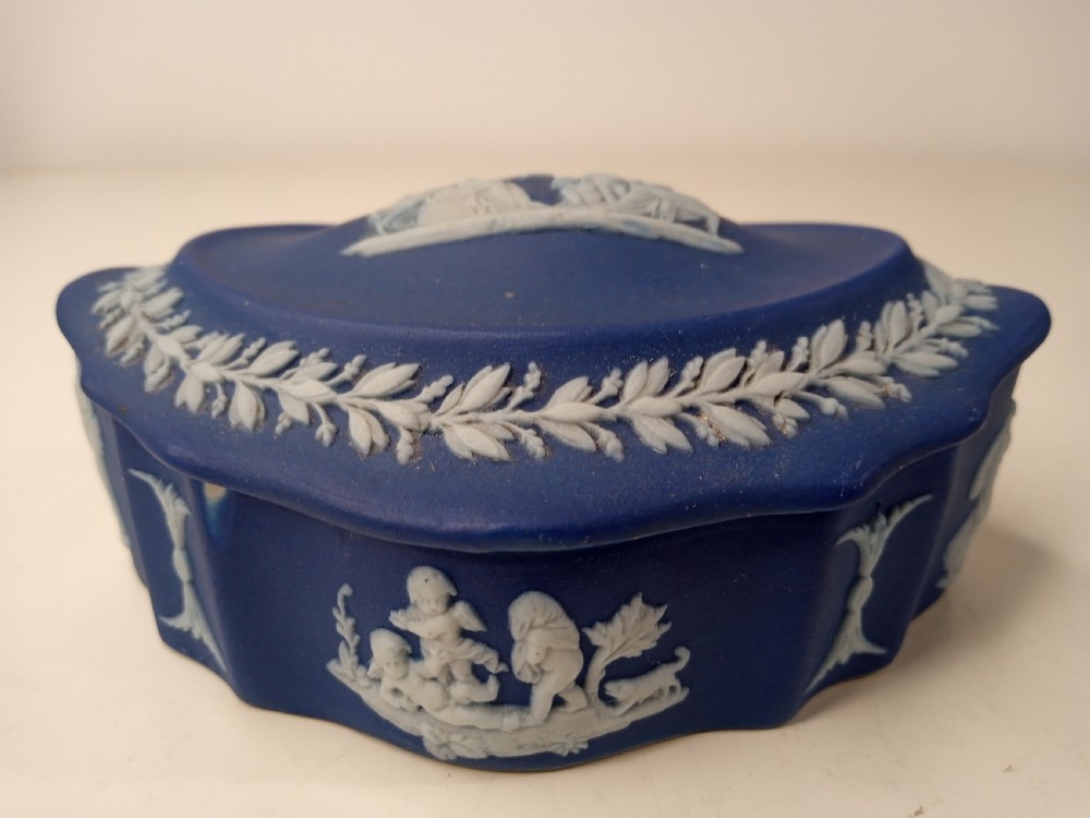 A dark blue oval jasperware lidded box featuring raised white classical bas-relief figures on the top surrounded by a white laurel leaf border. View V