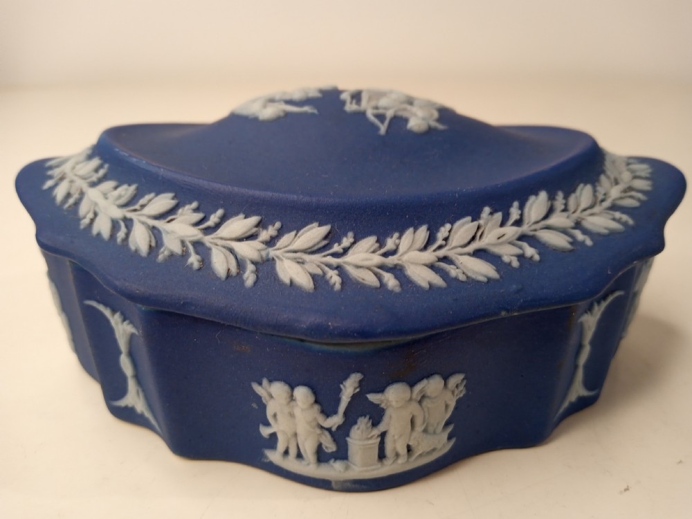 A dark blue oval jasperware lidded box featuring raised white classical bas-relief figures on the top surrounded by a white laurel leaf border. View VI