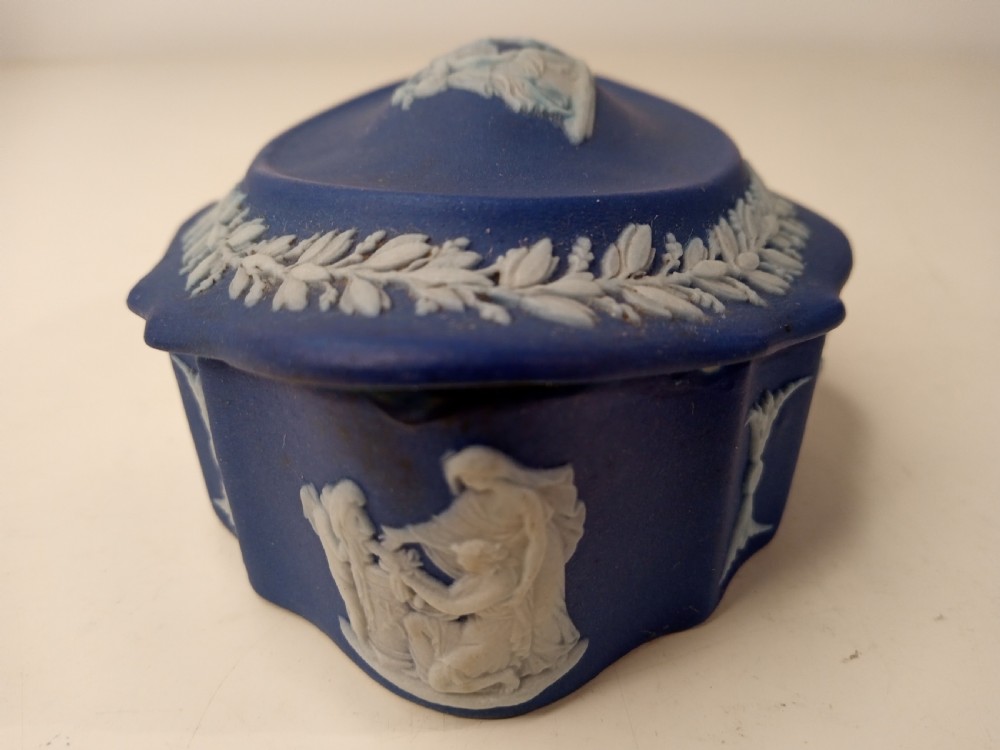 A dark blue oval jasperware lidded box featuring raised white classical bas-relief figures on the top surrounded by a white laurel leaf border. View VII