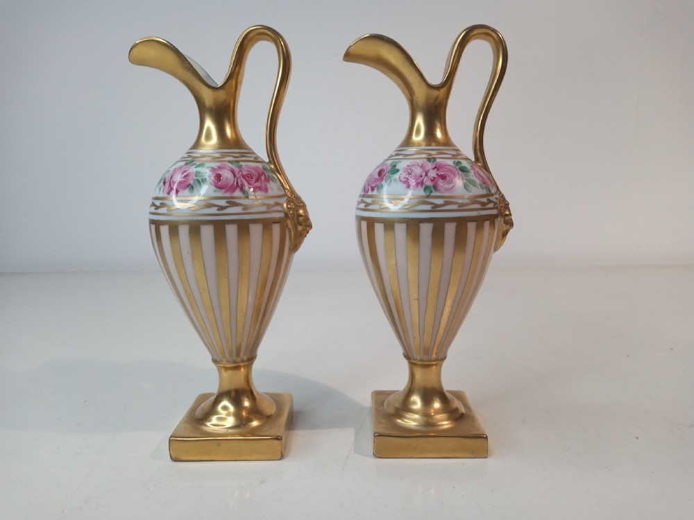 A pair of antique porcelain ewer vases featuring gilded spouts, vertical stripes, square pedestal bases, and hand-painted pink rose garlands around the shoulders.
