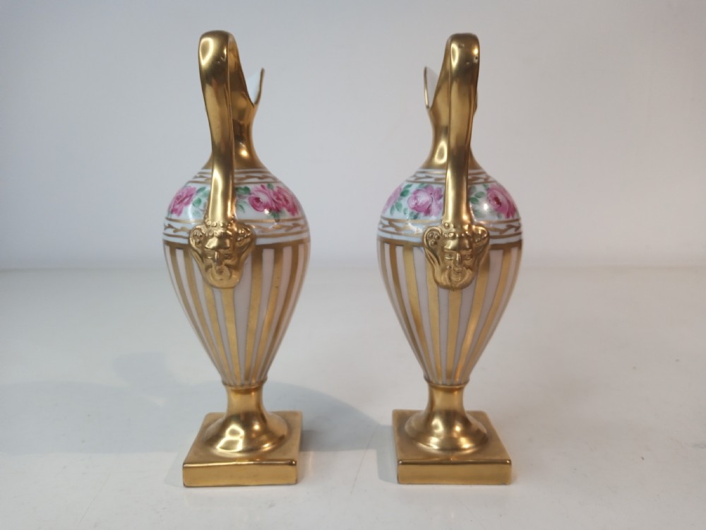 A pair of antique porcelain ewer vases featuring gilded spouts, vertical stripes, square pedestal bases, and hand-painted pink rose garlands around the shoulders. View II
