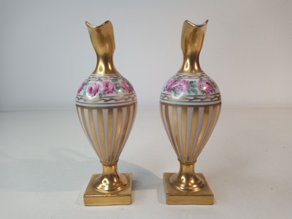 A pair of antique porcelain ewer vases featuring gilded spouts, vertical stripes, square pedestal bases, and hand-painted pink rose garlands around the shoulders. View IV