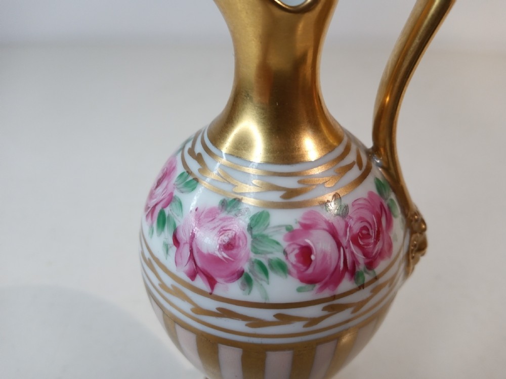 A pair of antique porcelain ewer vases featuring gilded spouts, vertical stripes, square pedestal bases, and hand-painted pink rose garlands around the shoulders. View VII