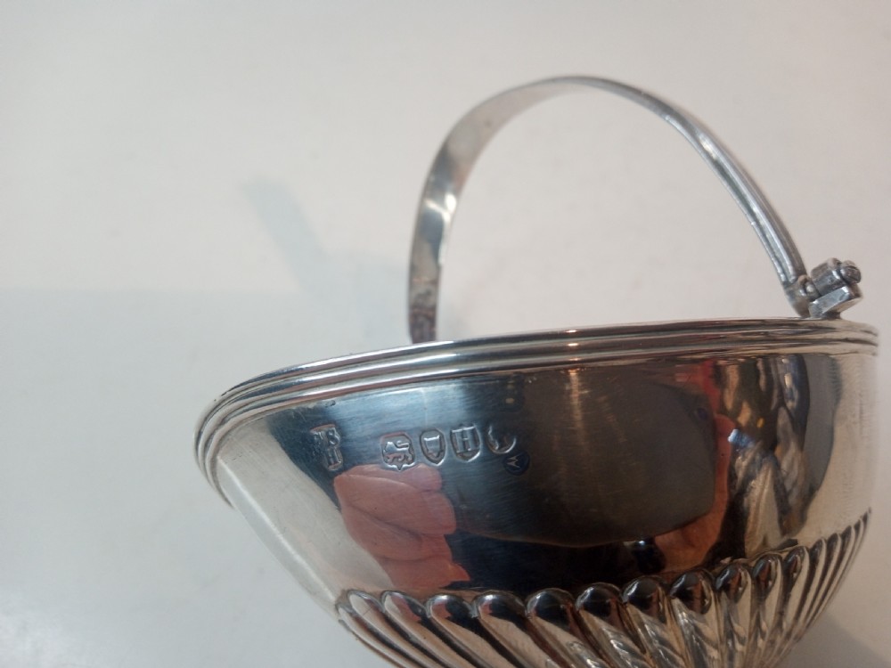 A Victorian sterling silver sweet basket featuring a pedestal foot, fluted detailing, and an upright swing handle. View VI