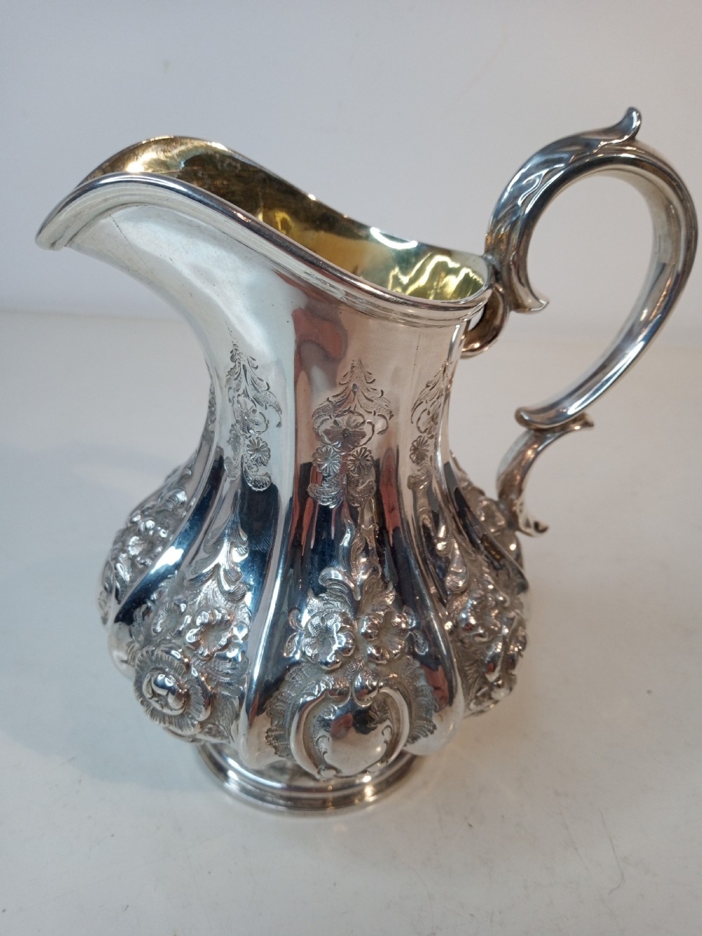 A Victorian sterling silver jug featuring hand-chased floral patterns, a fluted lower body, a scroll handle, and a gilt interior.