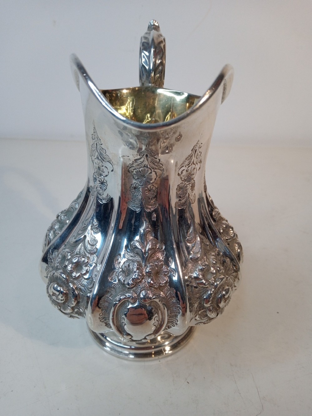 A Victorian sterling silver jug featuring hand-chased floral patterns, a fluted lower body, a scroll handle, and a gilt interior. View II