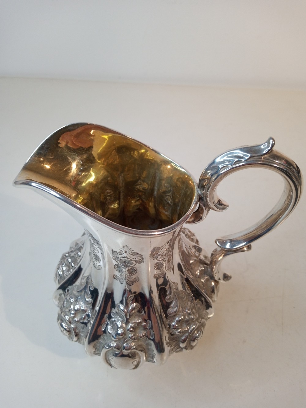 A Victorian sterling silver jug featuring hand-chased floral patterns, a fluted lower body, a scroll handle, and a gilt interior. View V