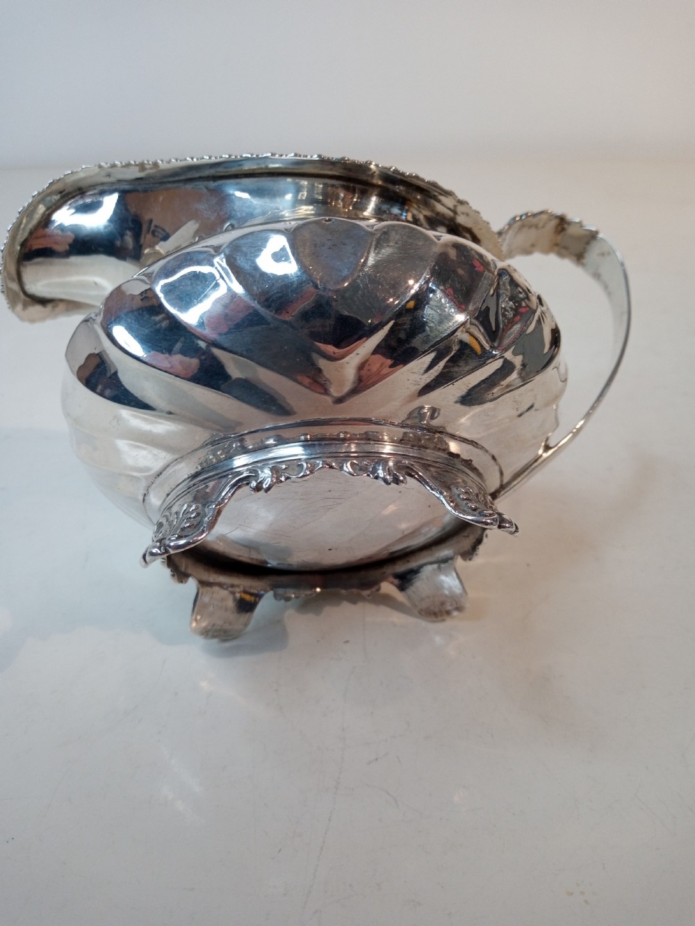 An antique Georgian sterling silver cream jug with a gadrooned rim, fluted lower body, cast handle, and four small feet. View V