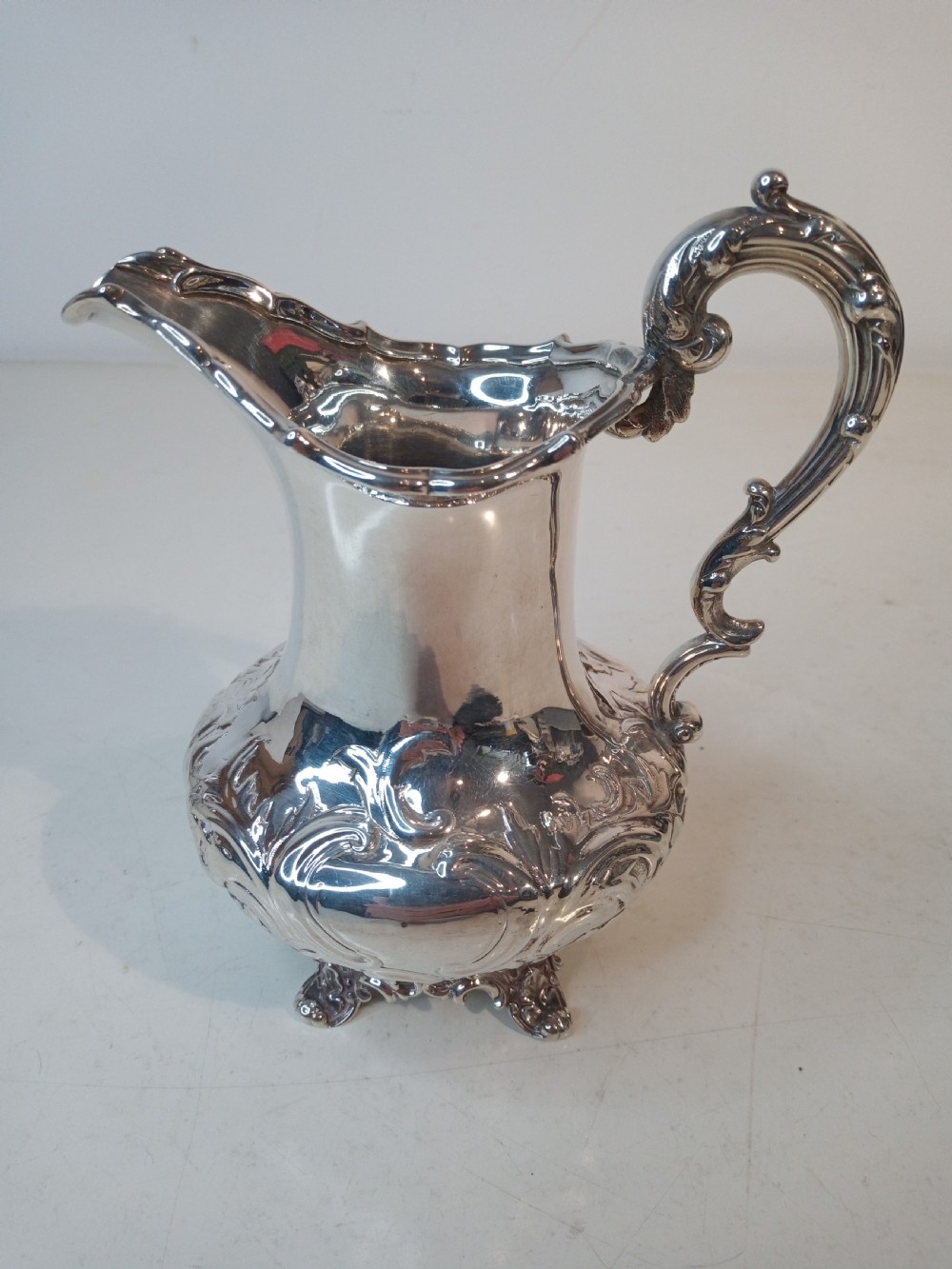 A Victorian hallmarked sterling silver jug with intricate scroll chasing, a shaped pour spout, a decorative handle, and scroll feet.